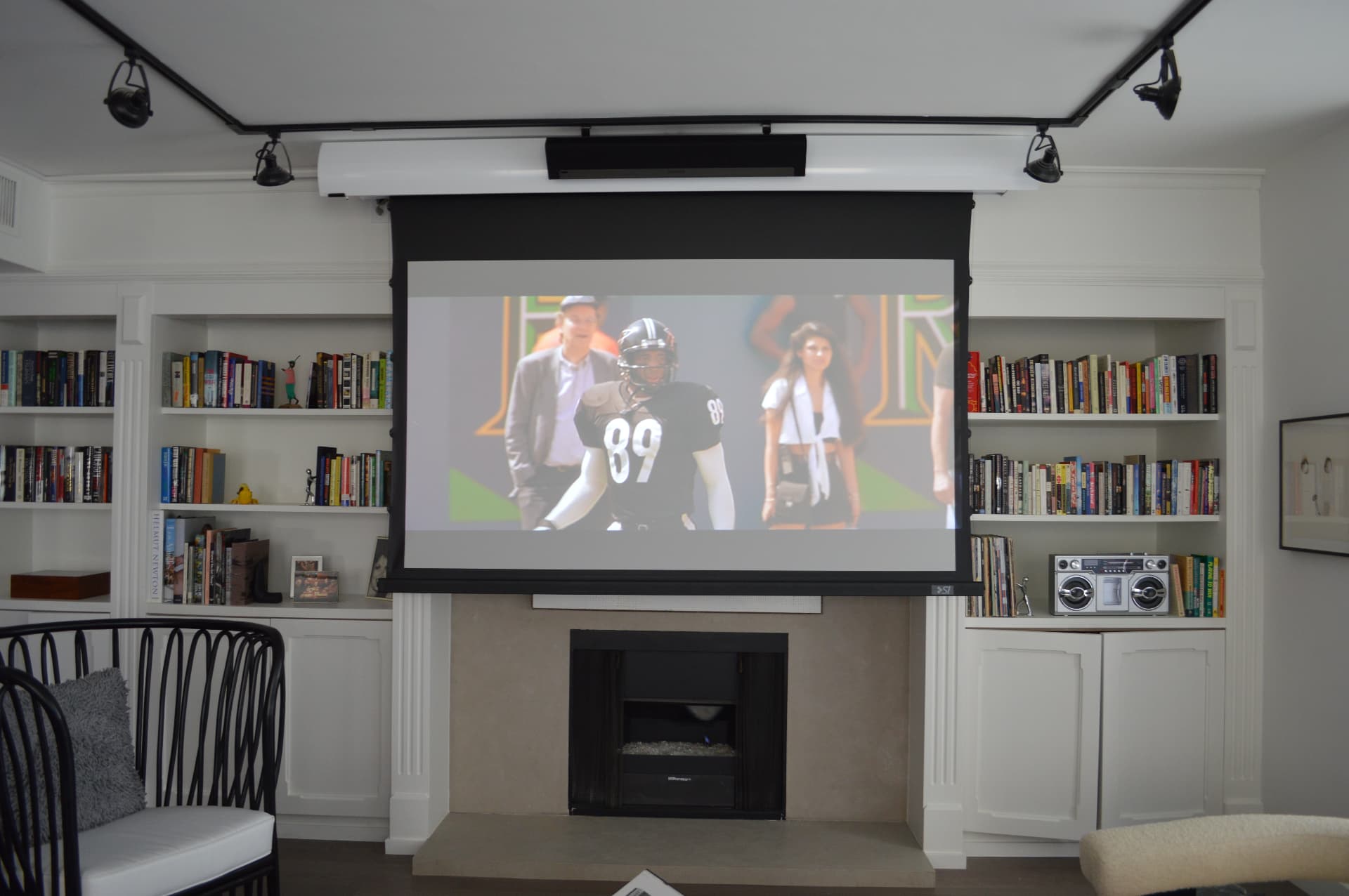 Motorized drop-down projection screen above fireplace displaying sports in elegant living room with built-in bookshelves and track lighting