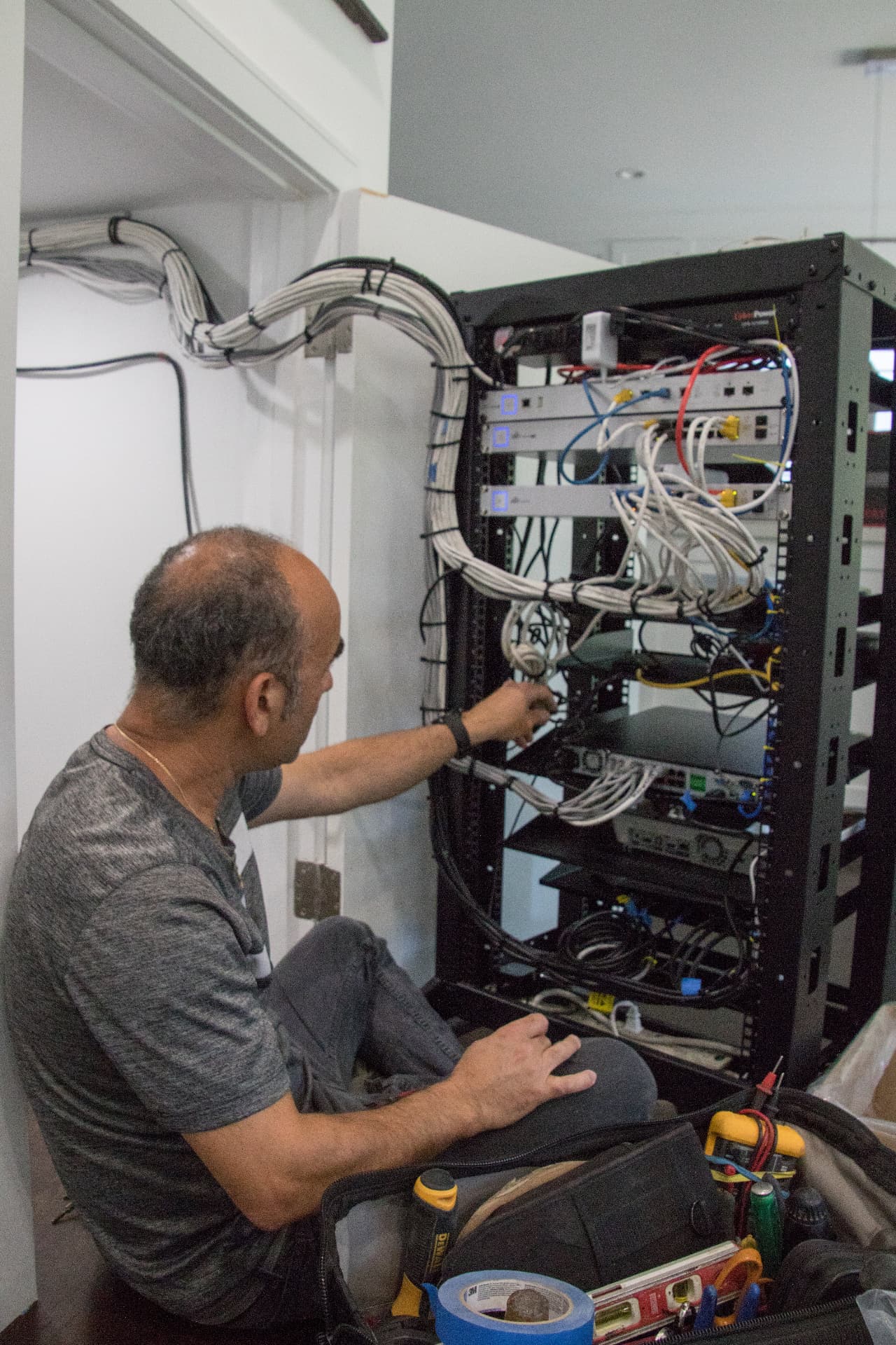 Technician Ron seated on the floor configuring a professional network rack with neatly dressed white data cables and equipment