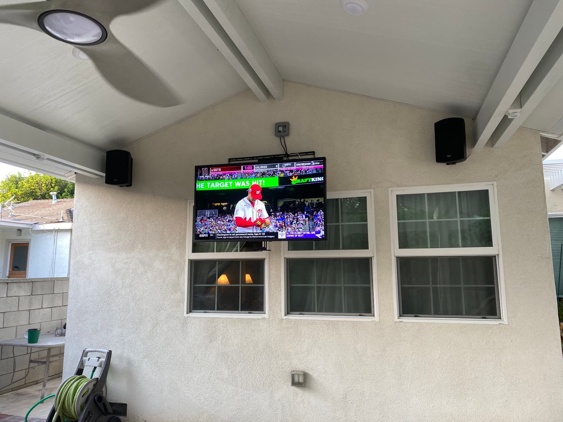 Large outdoor TV mounted under covered patio with flanking speakers, streaming live sports on a sunny day