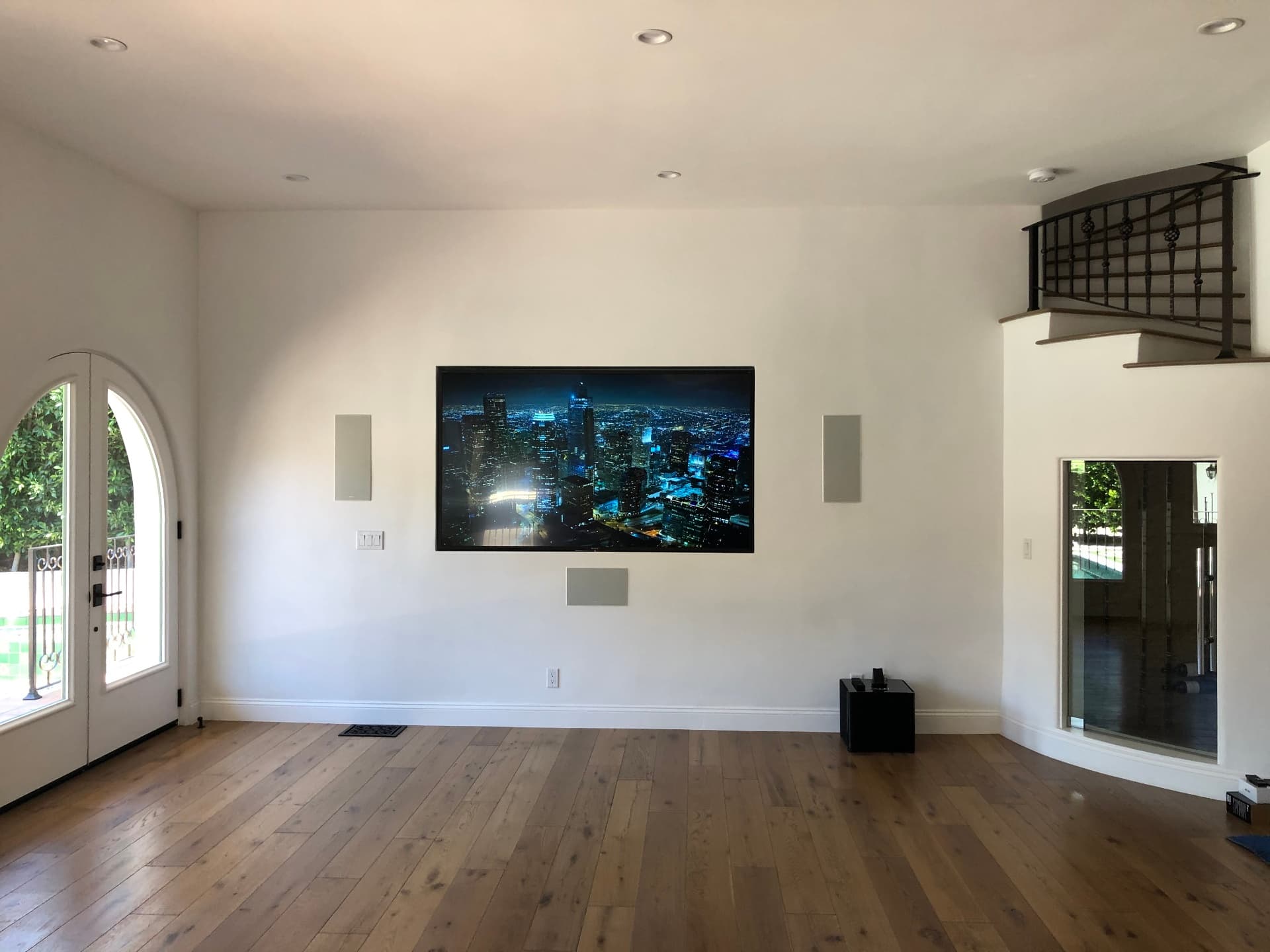 Large wall-mounted TV with in-wall surround speakers in Spanish-style living room featuring arched doorway, wrought iron balcony, and oak hardwood floors